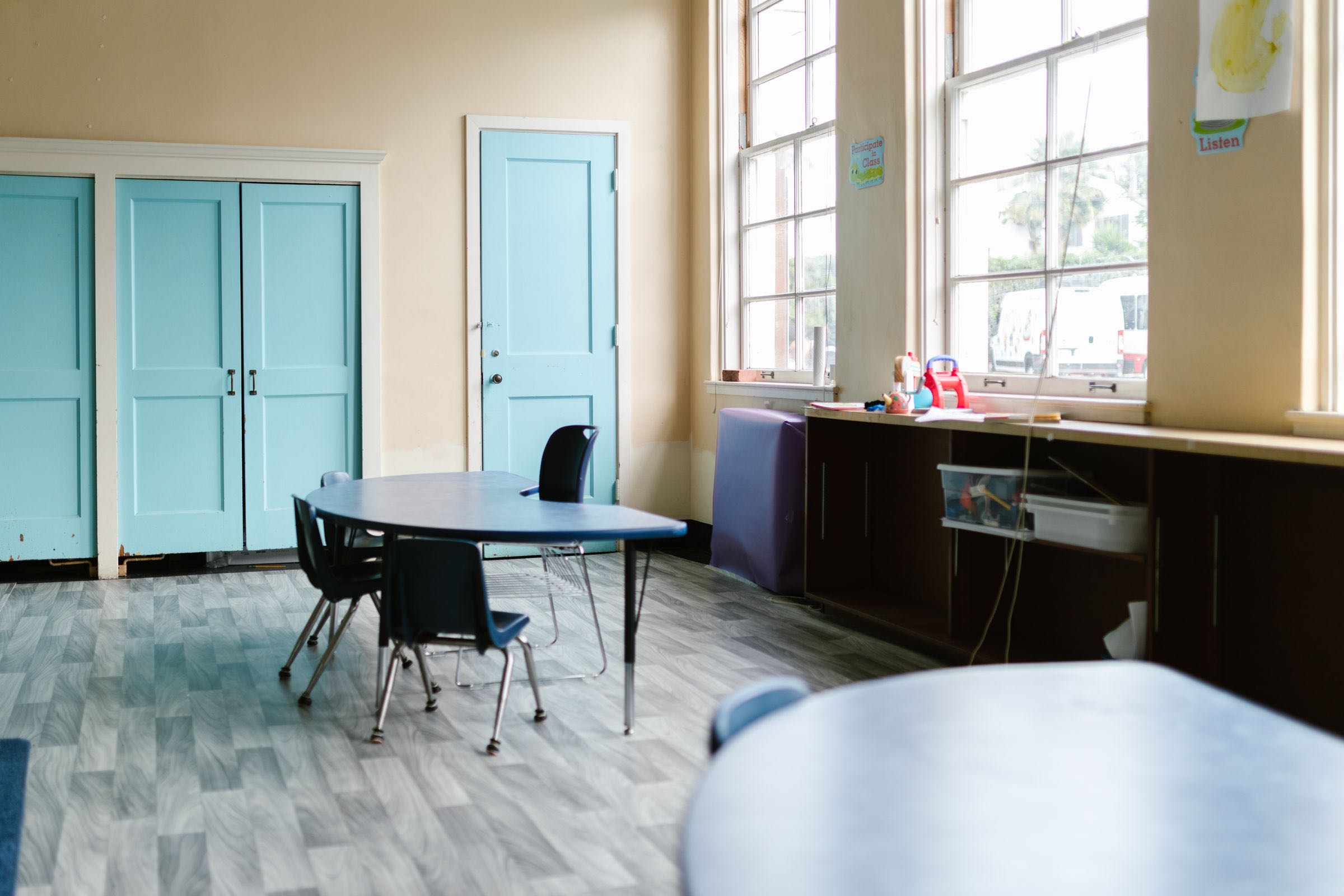 A sunlit classroom with tall windows, teal doors and a small round table with chairs.